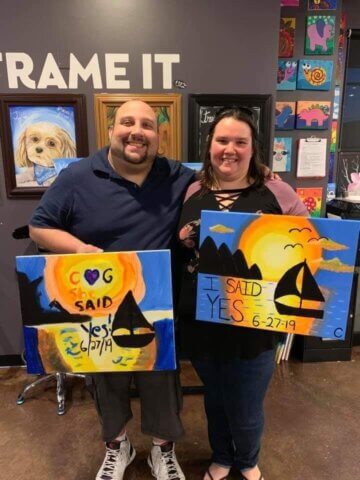 A couple smiling at the camera with two paintings in their hands. In his, a sunset with the words "she said yes." In hers, a sunset with the words "I said yes."