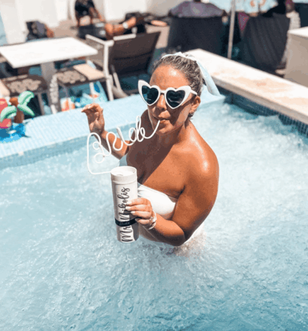 A bride in a hot tub wearing sunglasses and sipping a drink through a bride straw