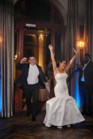 A bride with her hands in the air walking into a room with her groom