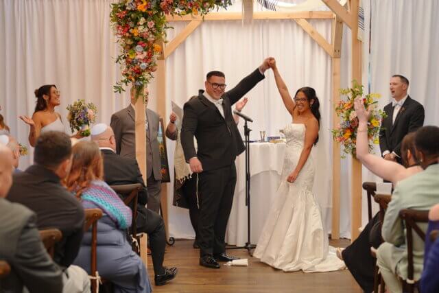 A bride and groom holding their hands in the air under a chuppah
